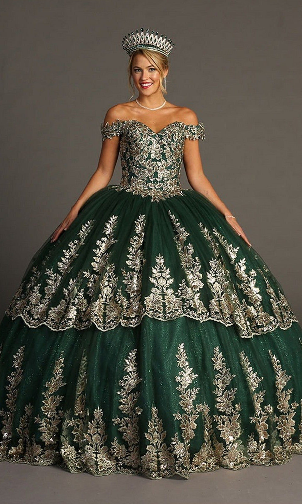 A woman stands against a plain background wearing the Quinceanera Dress CHF3083 By Chicasโa glitter tulle ballgown with gold embroidery, off-the-shoulder sleeves, and a matching crown.